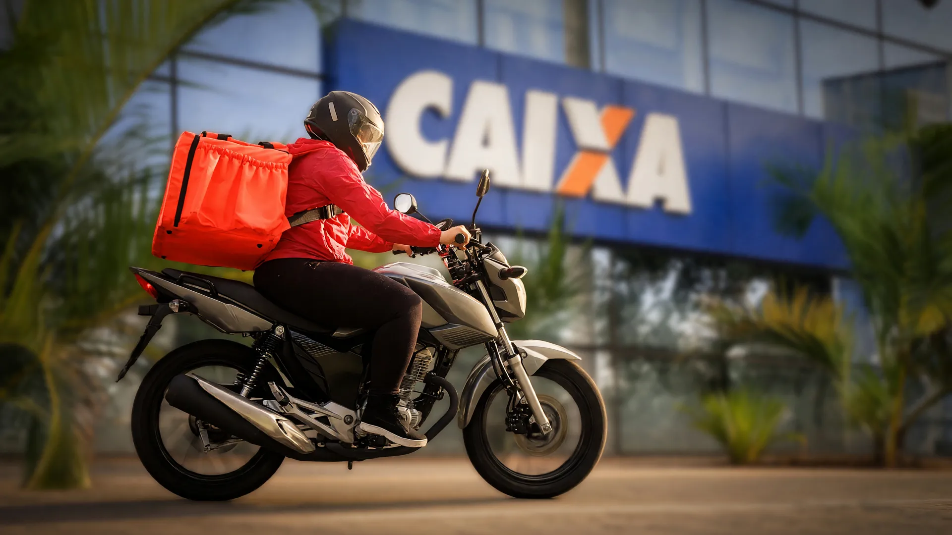 Entregador de app em sua moto, com agência da Caixa ao fundo, retratando o financiamento de moto para entregadores