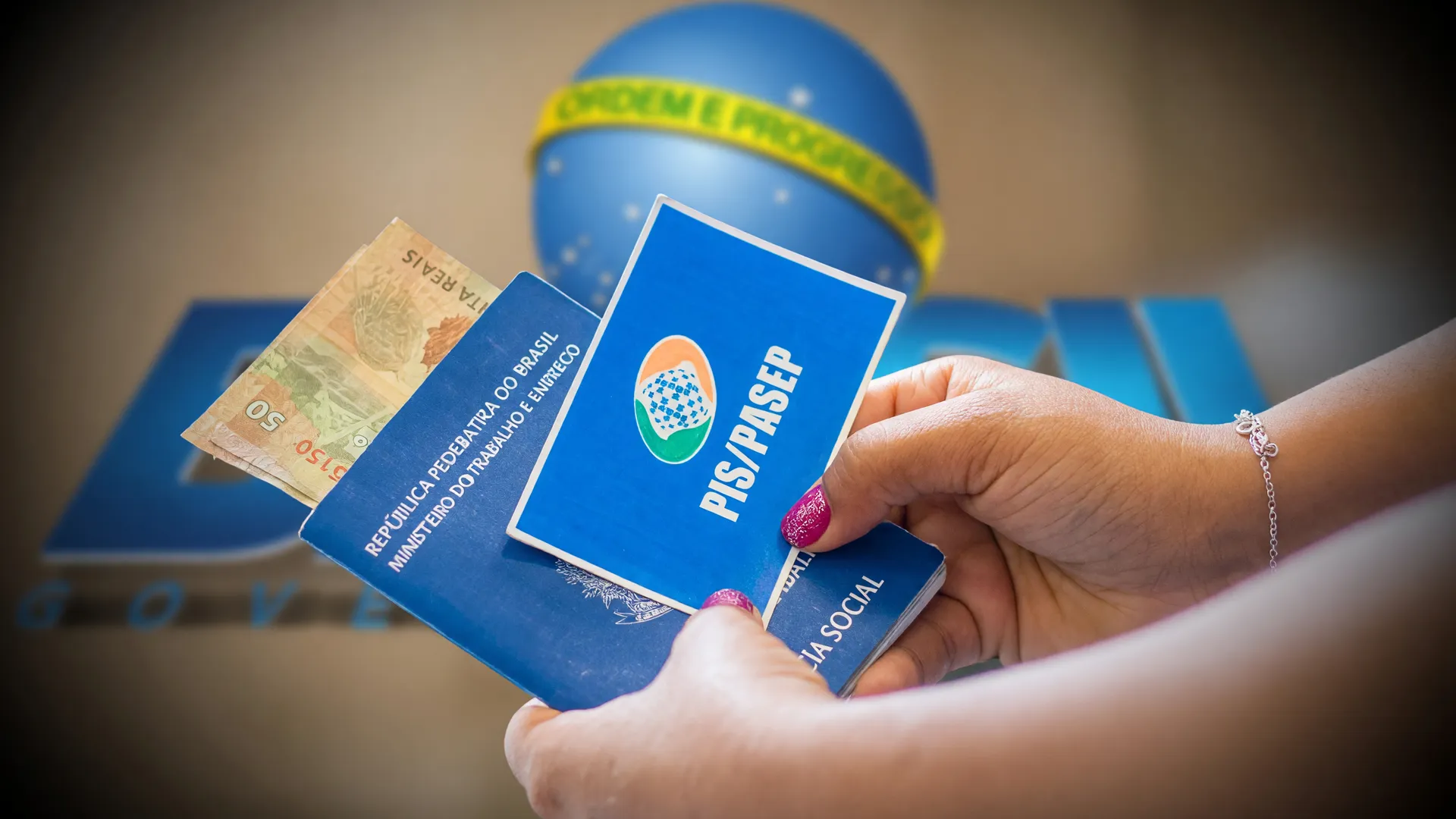 Mãos seguram cartões PIS/Pasep e cédula de real, representando o abono salarial PIS Pasep 2026